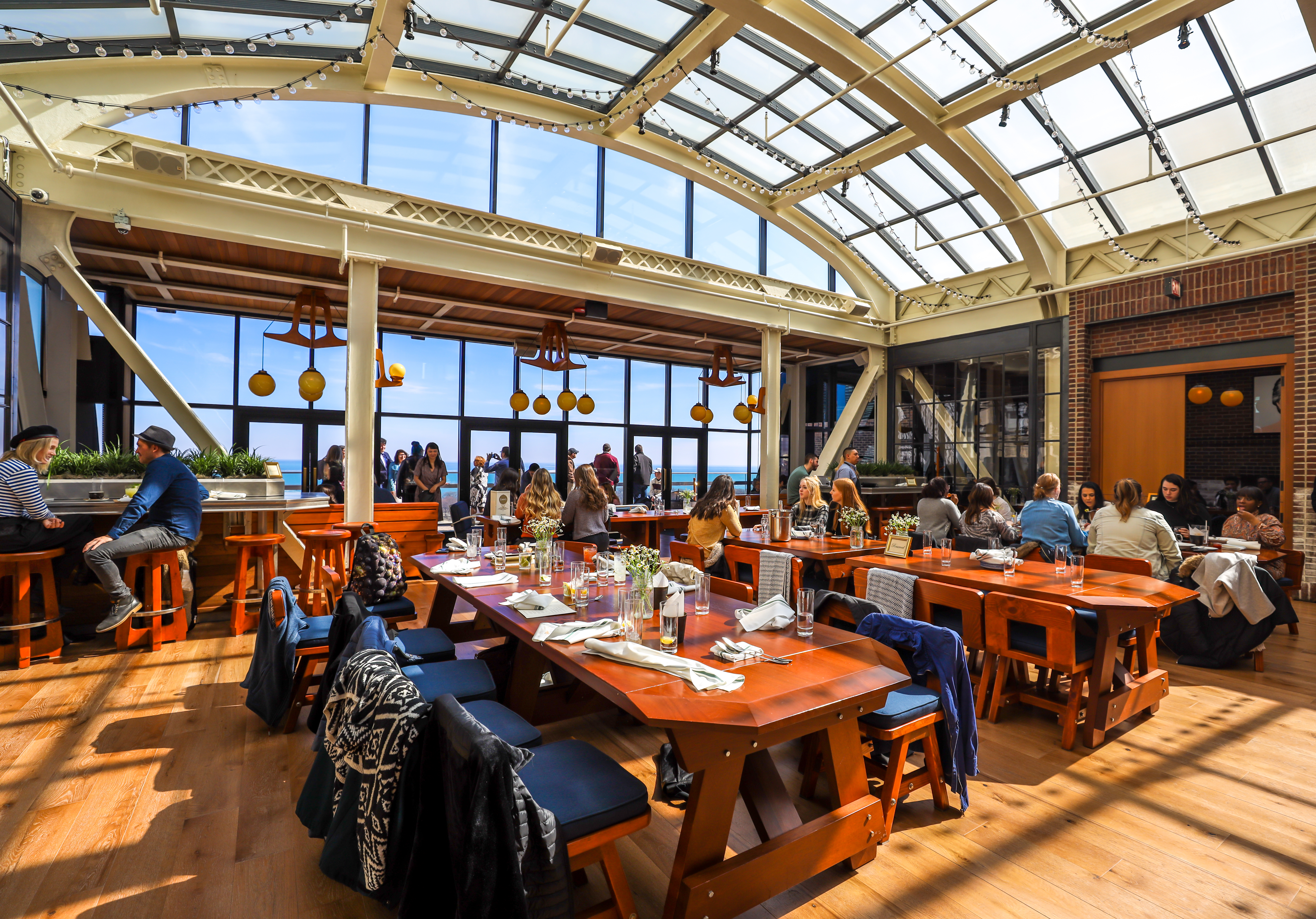 A large dining area with a glass roof and large windows, filled with people eating at wooden tables.