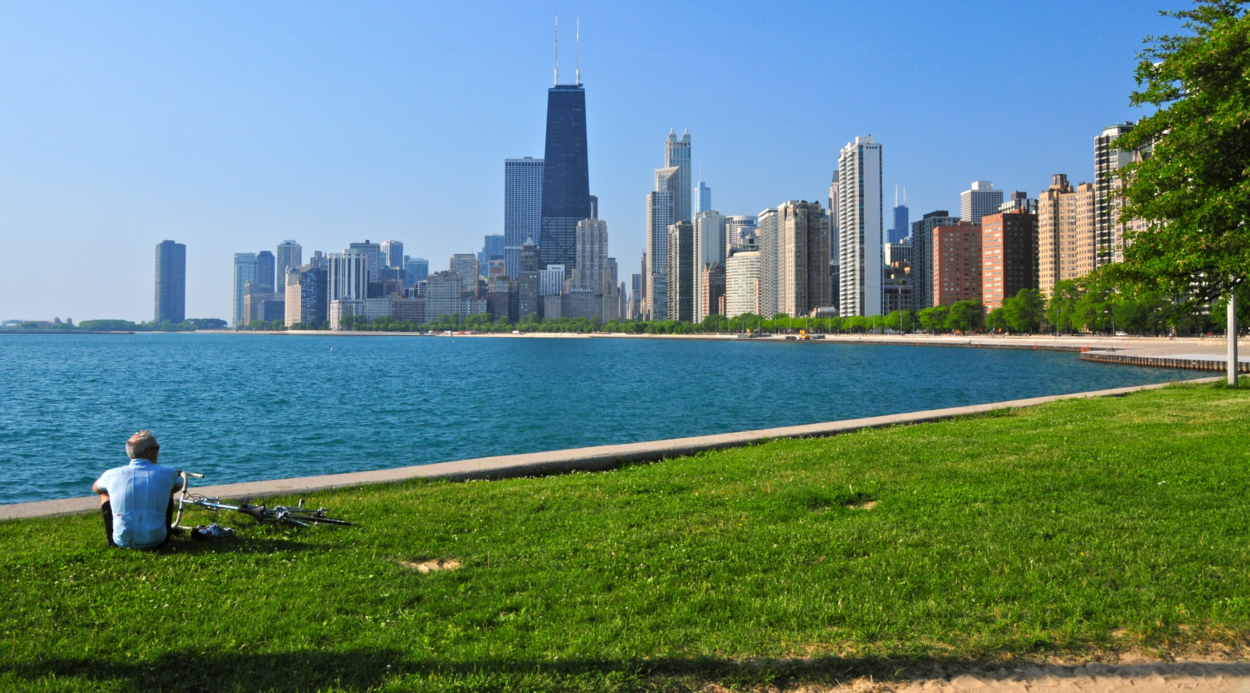 A cyclist sits on a vibrant green lawn, looking out over the blue waters of Lake Michigan toward the Chicago skyline on a clear, sunny day during spring in Chicago