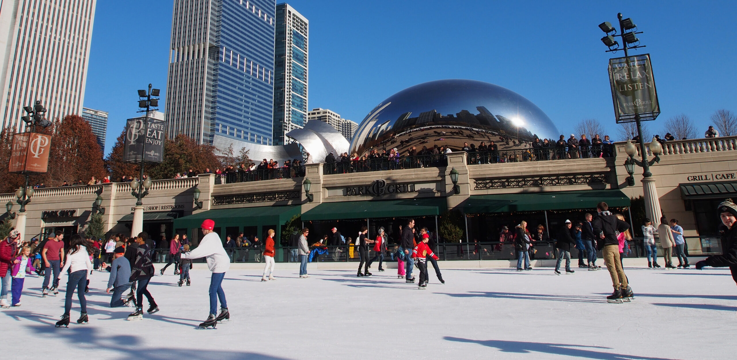 A group of people ice skating on the McCormick Tribune Ice Rink in Millennium Park, Chicago, on a bright, sunny day.