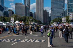Crowds fill a downtown Chicago street during the Taste of Chicago food festival, with festival tents, city skyscrapers, and event banners lining the avenue on a sunny day.