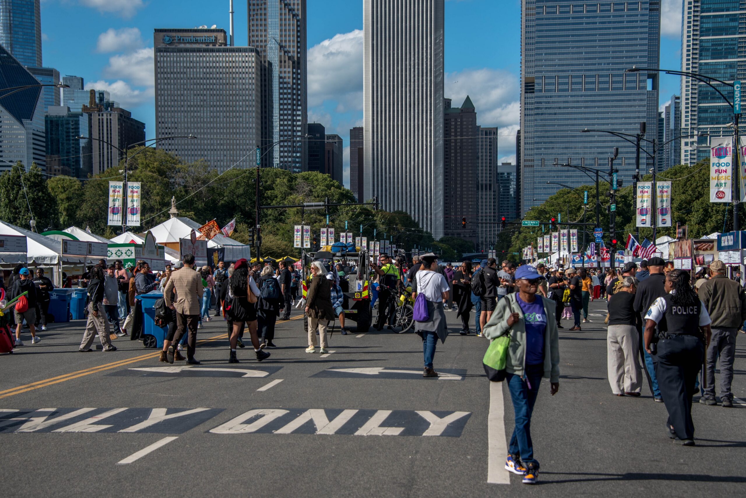 Crowds fill a downtown Chicago street during the Taste of Chicago food festival, with festival tents, city skyscrapers, and event banners lining the avenue on a sunny day.