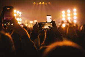 A crowd at a nighttime concert holds up smartphones to record a brightly lit stage, with warm golden lights and blurred silhouettes creating a lively live music atmosphere.
