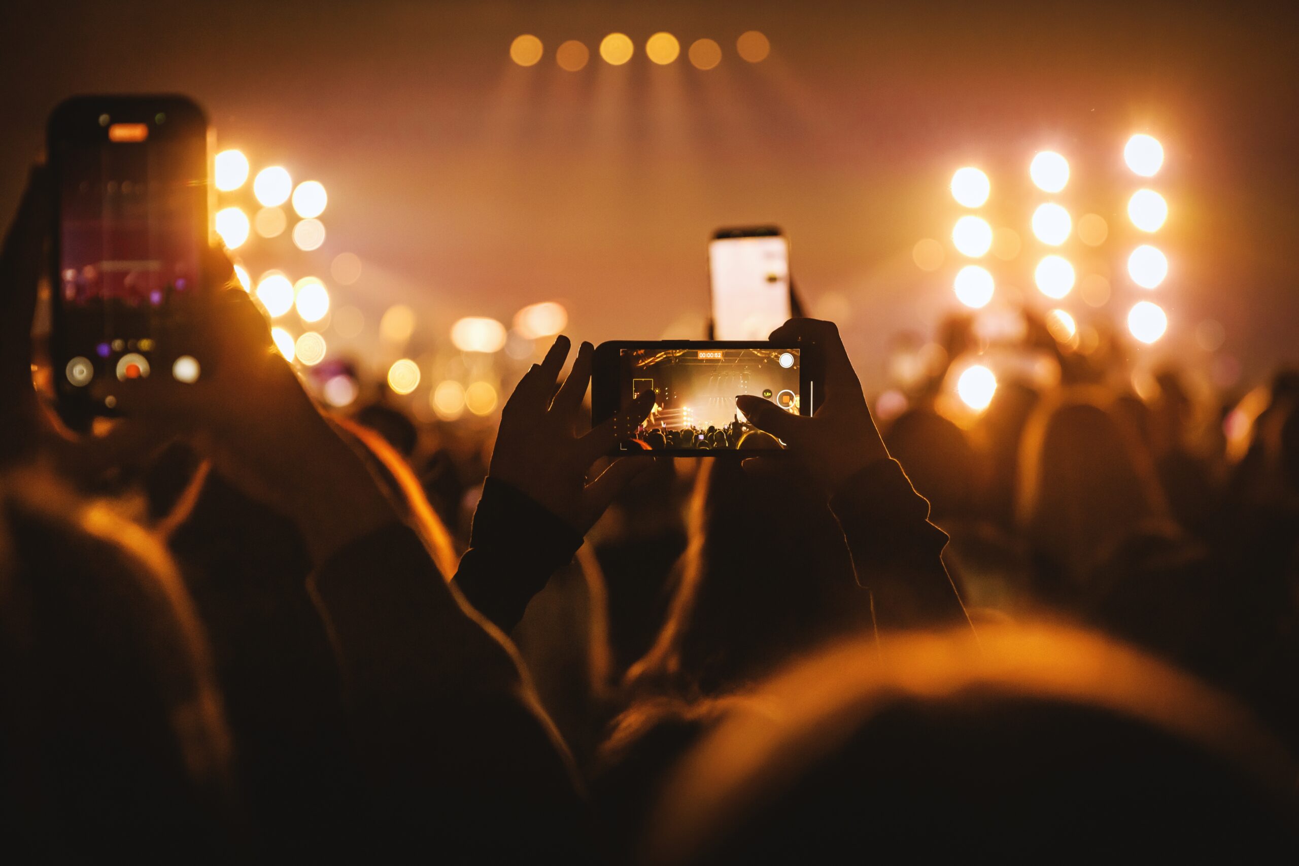 A crowd at a nighttime concert holds up smartphones to record a brightly lit stage, with warm golden lights and blurred silhouettes creating a lively live music atmosphere.