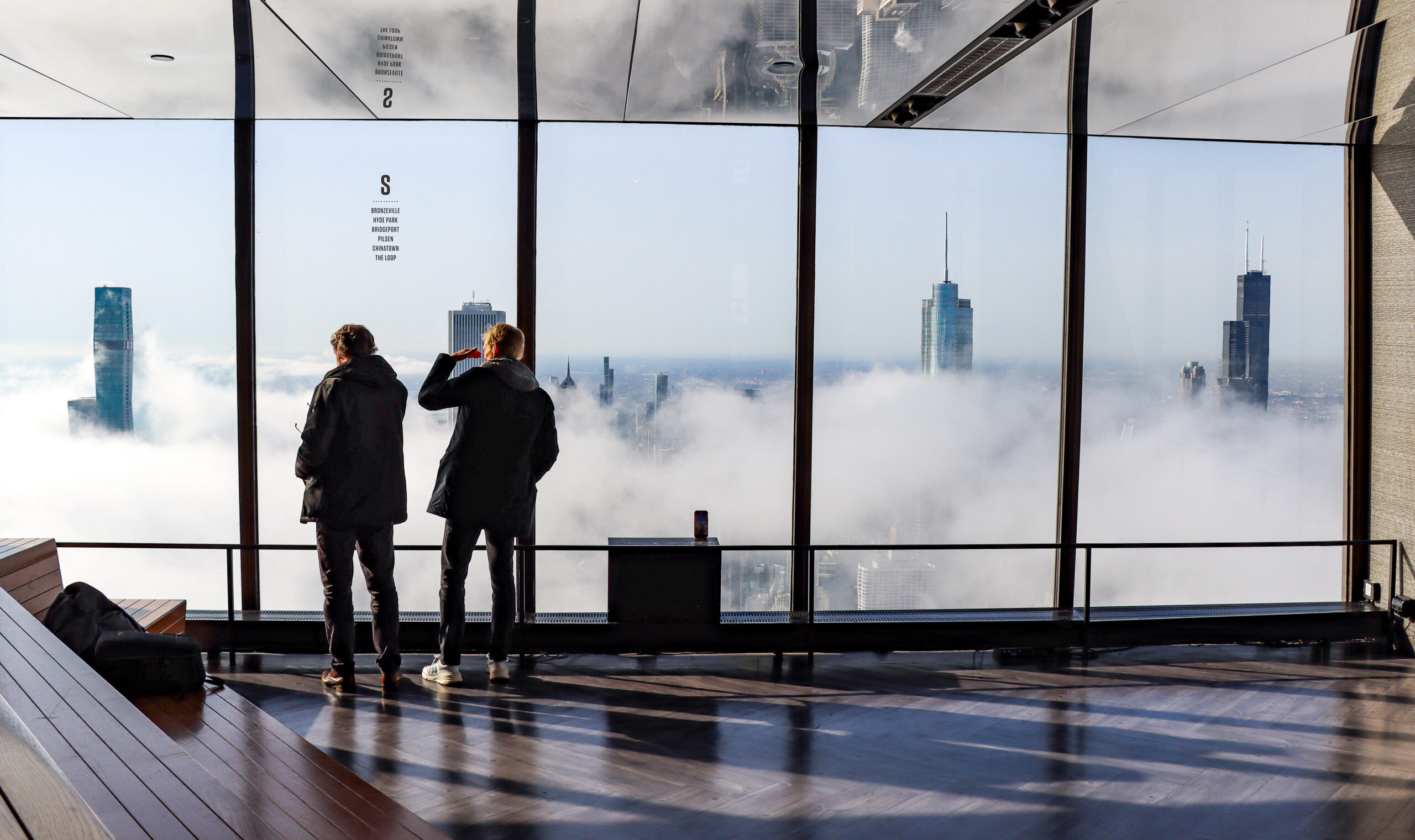 Two people looking out at a city skyline through large windows with clouds below.