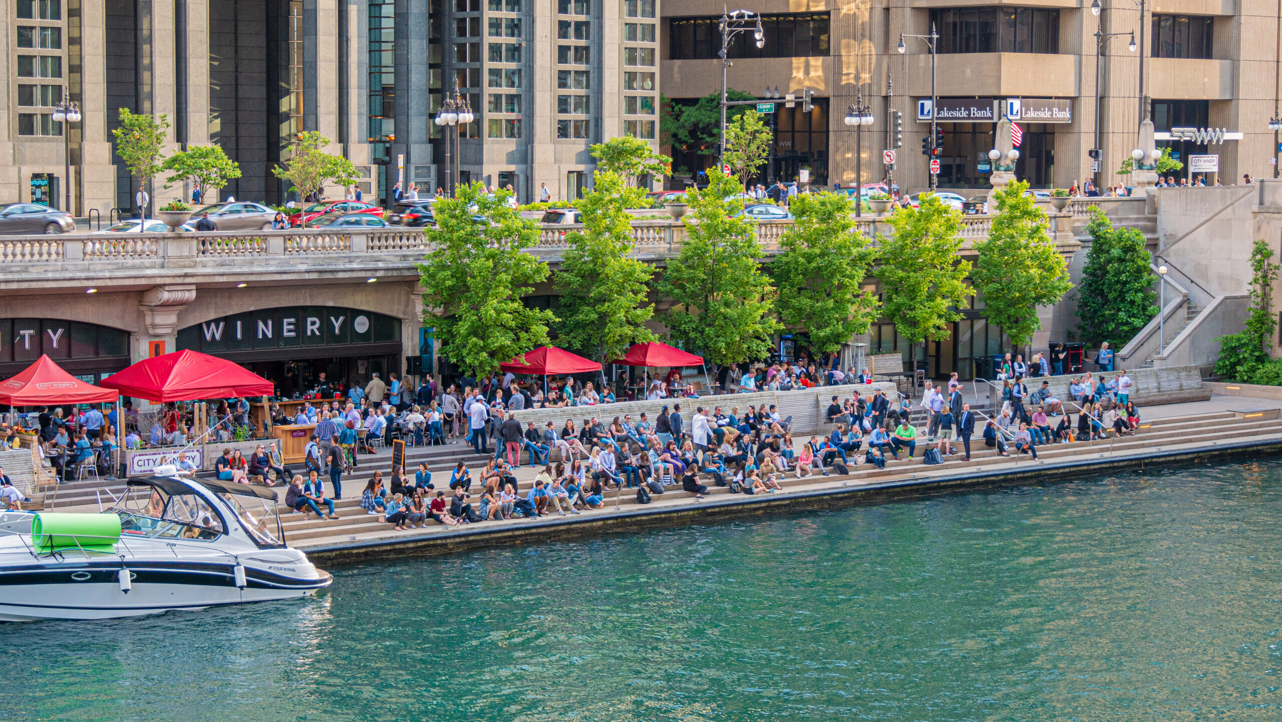 Chicago Riverwalk with people dining and relaxing along the river, surrounded by downtown buildings, trees, and boats.