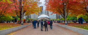 Cloud Gate ("The Bean") in Chicago's Millennium Park, framed by a path lined with red and orange fall foliage as visitors walk toward it, with the city skyline behind.