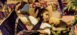 A young dancer in a flower-trimmed straw hat and a white lace blouse swirls a purple floral skirt while performing in a nighttime Halloween parade in Chicago, with spectators watching in the background.