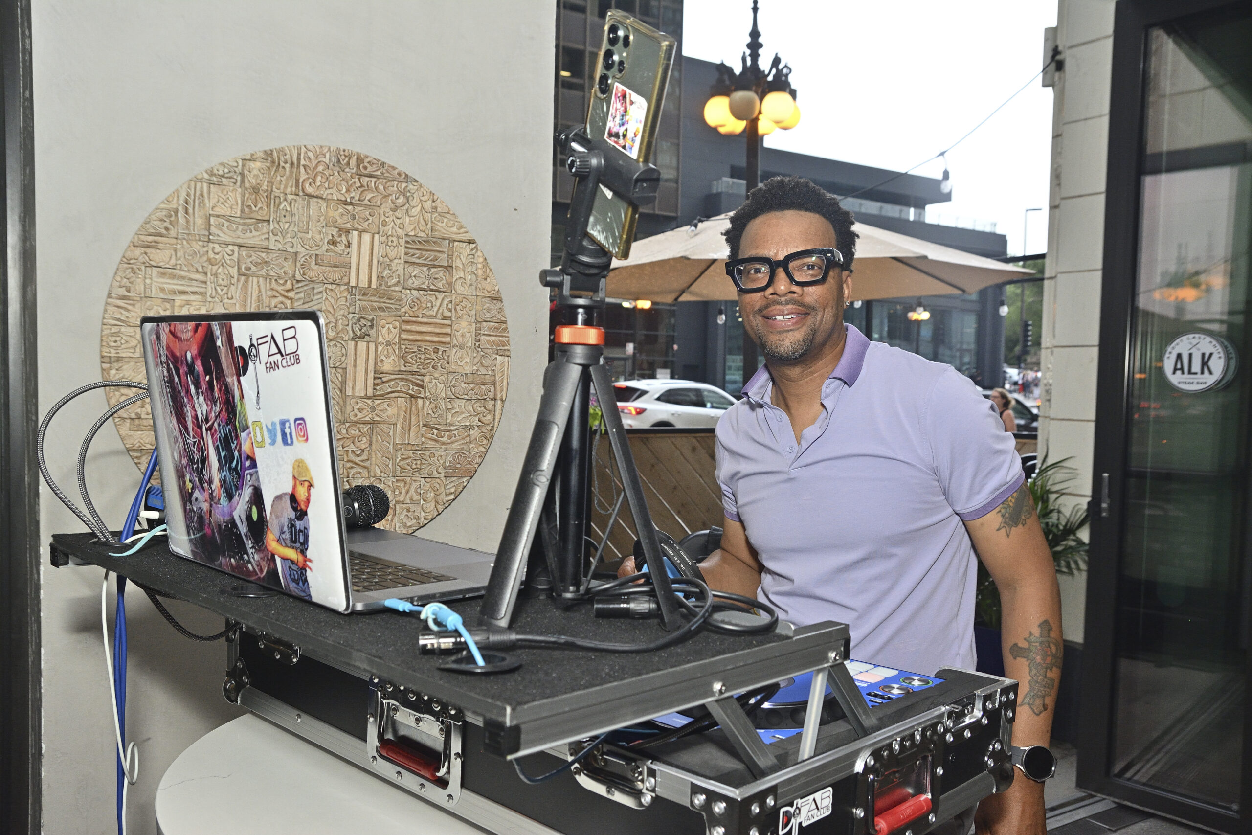 A person wearing glasses and a light purple shirt, standing behind a DJ setup with a laptop, camera on tripod, and other equipment, with an outdoor background.