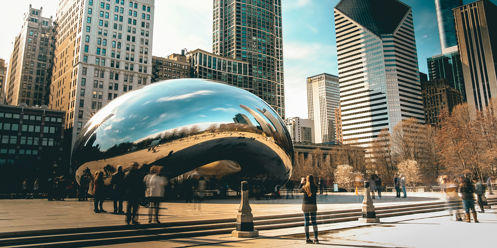 People stand around the reflective Cloud Gate sculpture in Millennium Park, Chicago, with surrounding high-rise buildings visible.