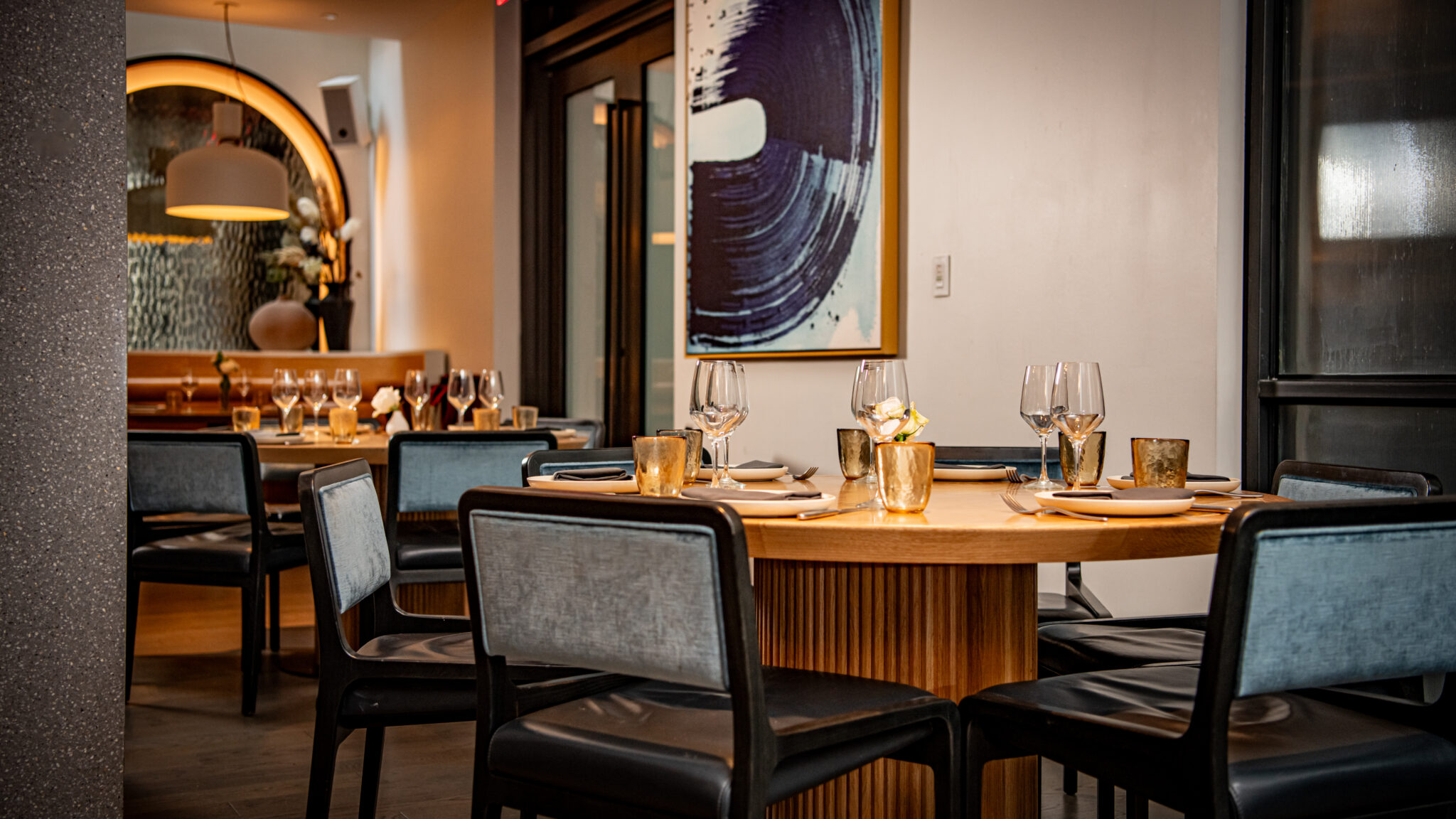 A dining area with a round wooden table set with wine glasses, plates, and candles, surrounded by blue chairs.