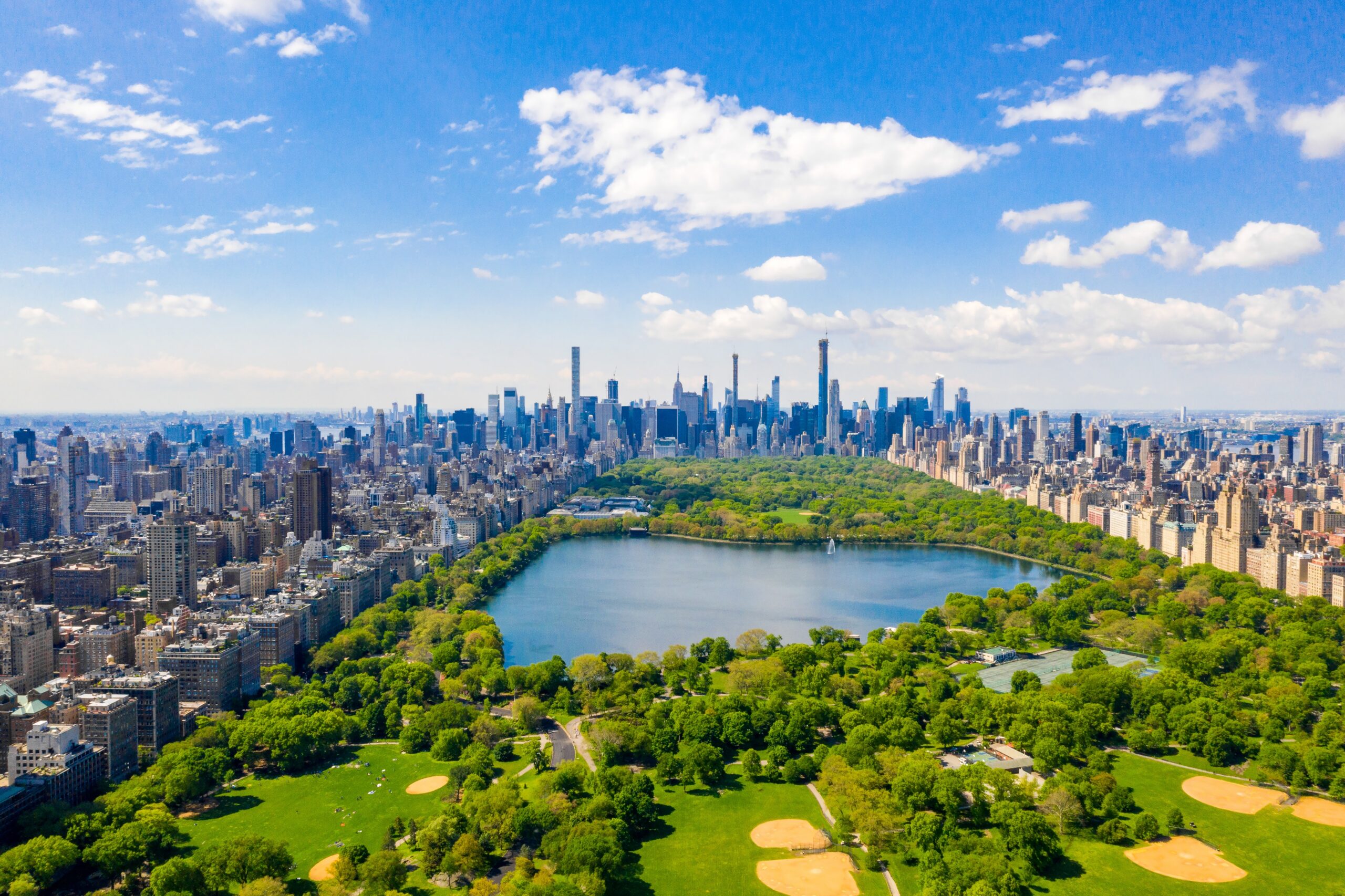 An aerial, high-angle view of Central Park in New York City on a bright, sunny day. The lush green landscape of the park, featuring the Jacqueline Kennedy Onassis Reservoir and several baseball diamonds, is framed by the dense, towering skyscrapers of the Manhattan skyline under a blue sky with scattered white clouds.