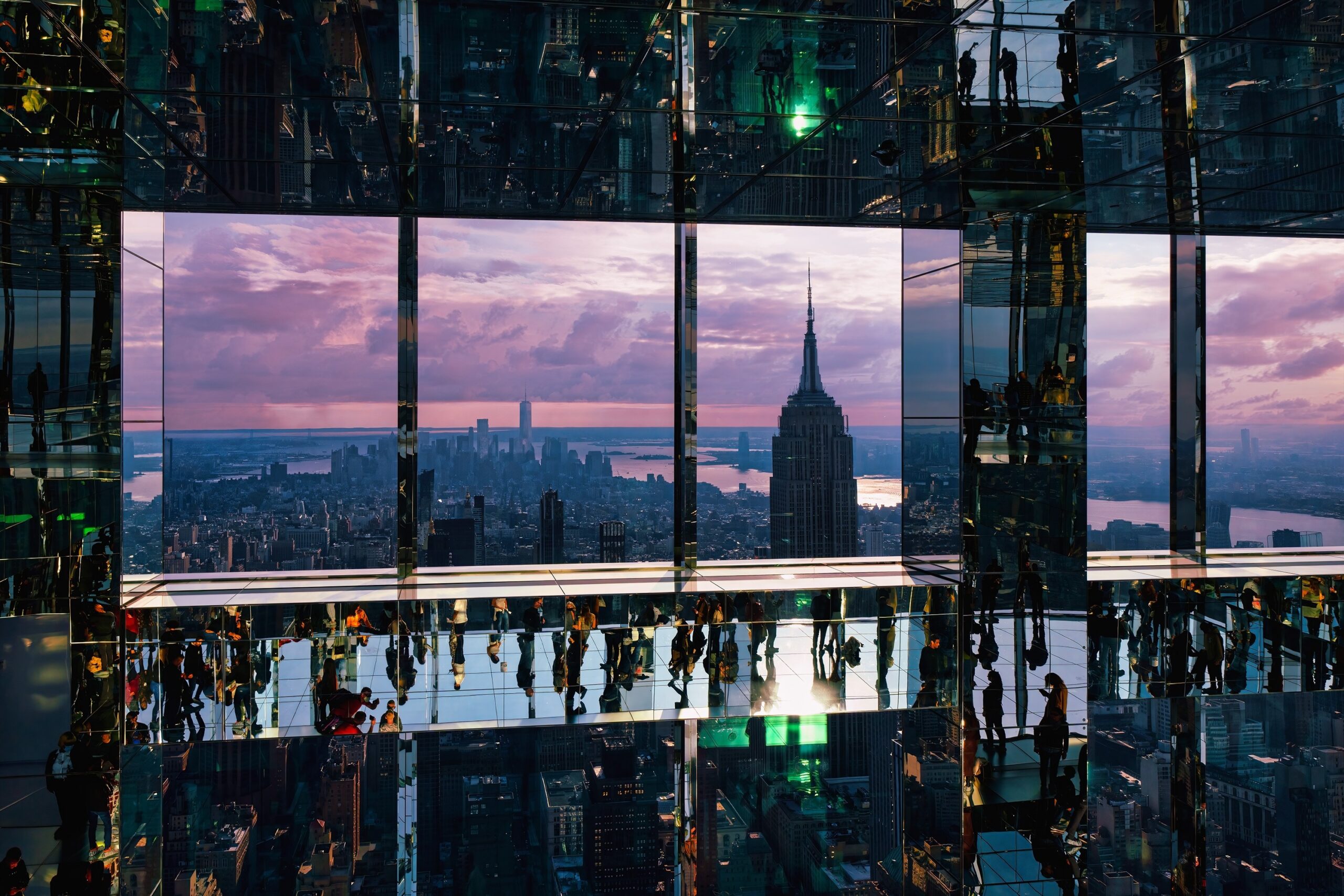 A high-angle, wide shot of the Summit One Vanderbilt observation deck in New York City during sunset. The room is a kaleidoscopic hall of mirrors with reflective floors, walls, and ceilings, creating an infinite, surreal environment.