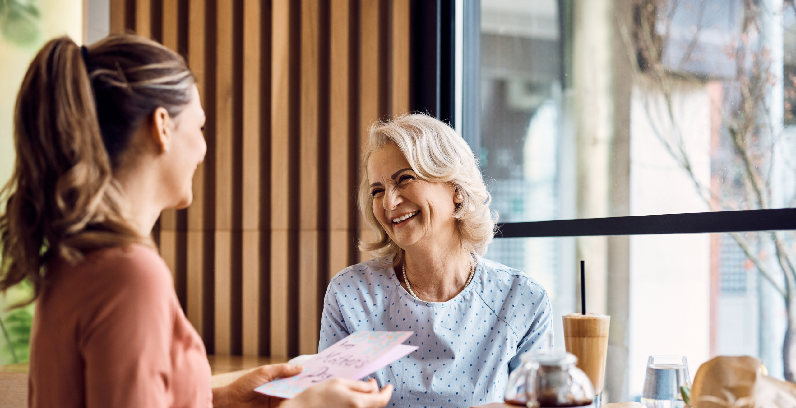 A daughter hands a Mother's Day card to her smiling mother in a modern, stylish restaurant setting with warm wood accents and large windows.