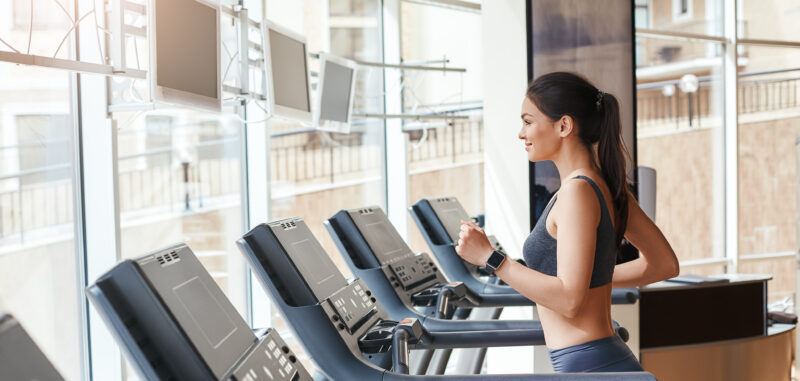 Woman jogging on a treadmill in a modern gym with large windows and multiple cardio machines.