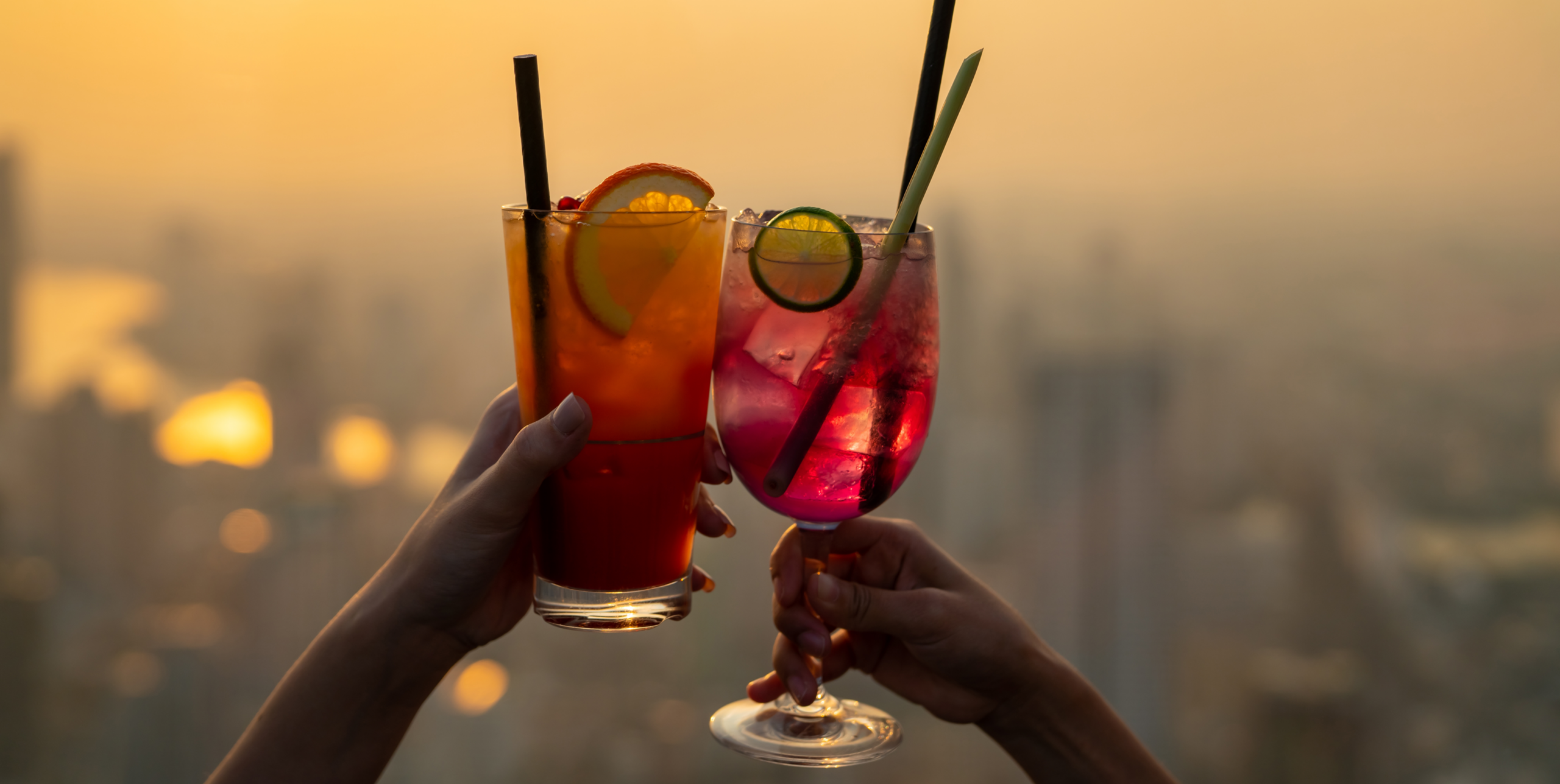 Two people raise colorful cocktails for a toast at a rooftop bar during sunset, with a blurred city skyline in the background and citrus garnishes adding a vibrant touch.
