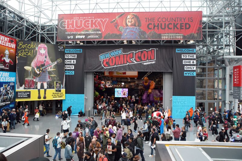 Crowds of fans gather at the entrance to New York Comic Con at the Javits Center, surrounded by Comic Con banners, character displays, and event signage.