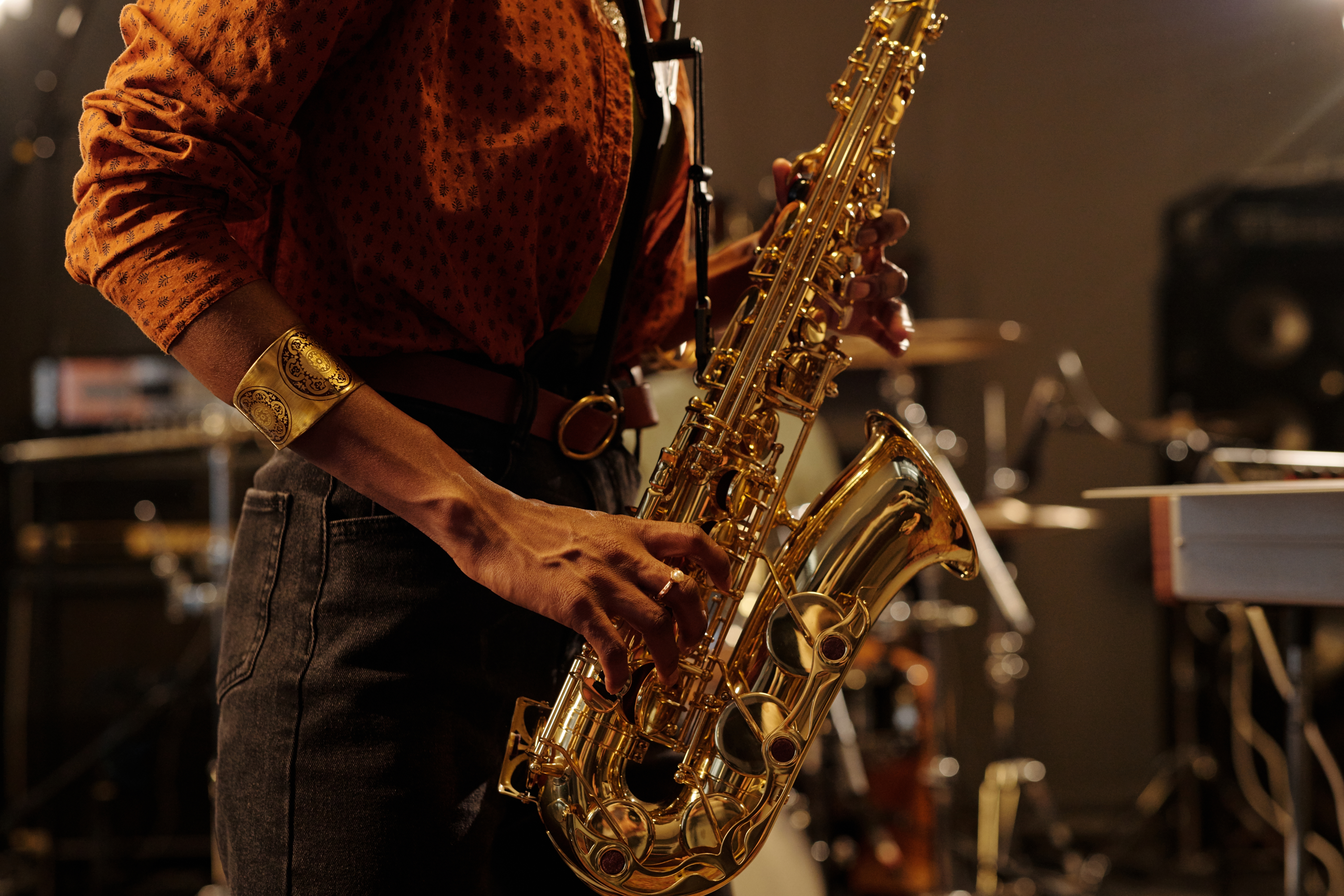 Close-up of a saxophonist's hands playing a gold saxophone on a dimly lit stage, with drum and speaker equipment blurred in the background.