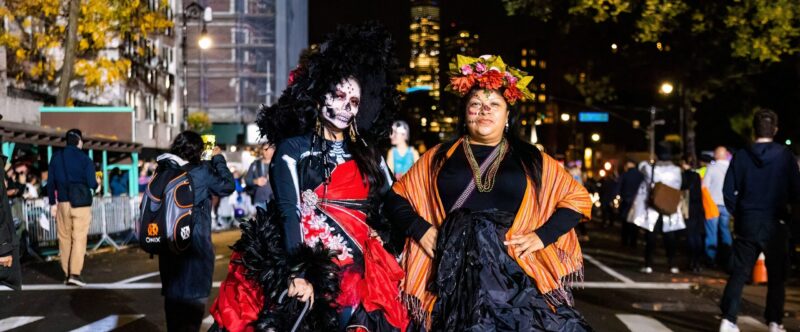 Two people in Day of the Dead costumes with face paint, flowers, and ruffled gowns pose on a lit NYC street on Halloween night.