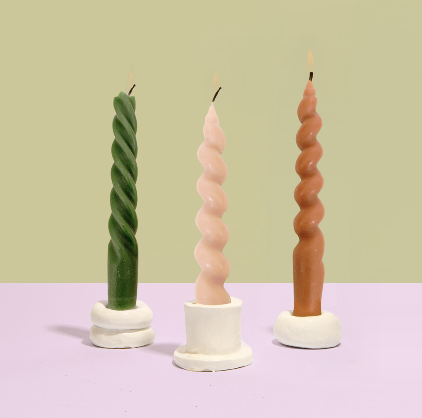 Three twisted candles in green, pink, and brown are placed in white holders, set against a pastel background.