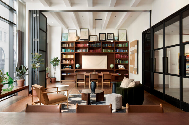 A photograph of a mid-century modern room in Arlo NoMad, showcasing **NoMad hotel meeting spaces**, with leather and green velvet armchairs, a rug, a bookshelf wall, and text overlay.