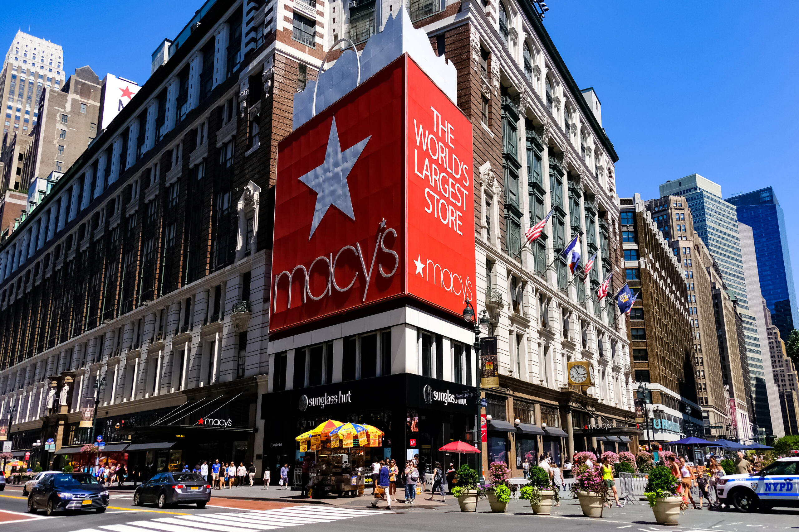 A street-level corner view of the historic Macy's flagship store at Herald Square in New York City, featuring its massive red "World's Largest Store" star signage and classic stone architecture under a clear sky.