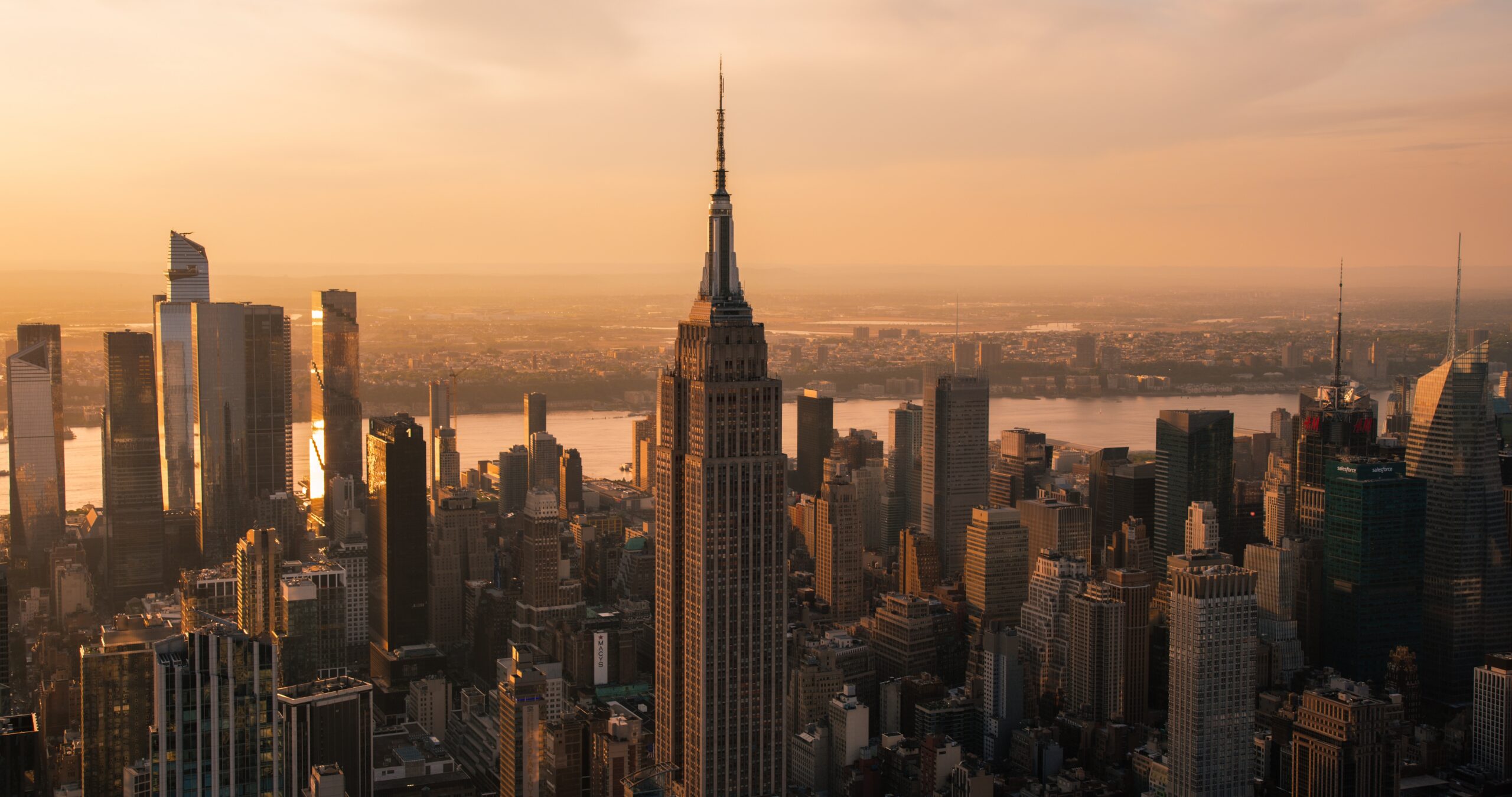 A high-angle aerial view of the New York City skyline at sunset, featuring the Empire State Building prominently in the center with the Hudson River and Hudson Yards in the background under a warm orange glow.