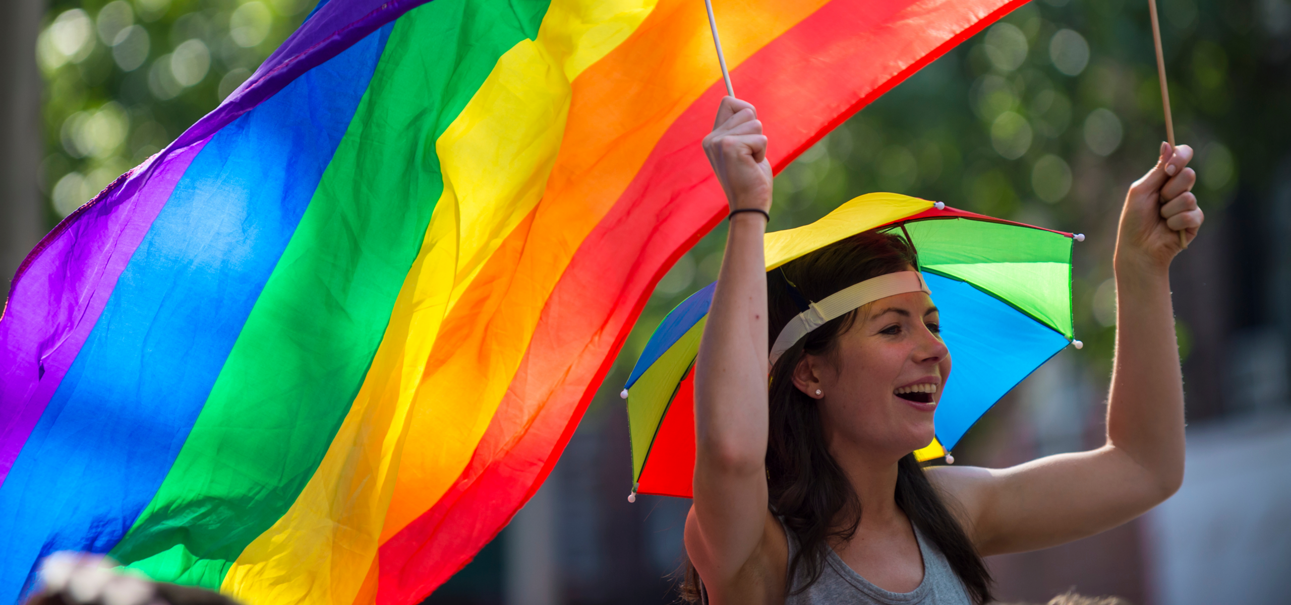 A smiling person at a pride event wears a rainbow umbrella hat and holds up a large, waving rainbow flag alongside two small American flags. The background is a soft-focus view of green trees in bright daylight.
