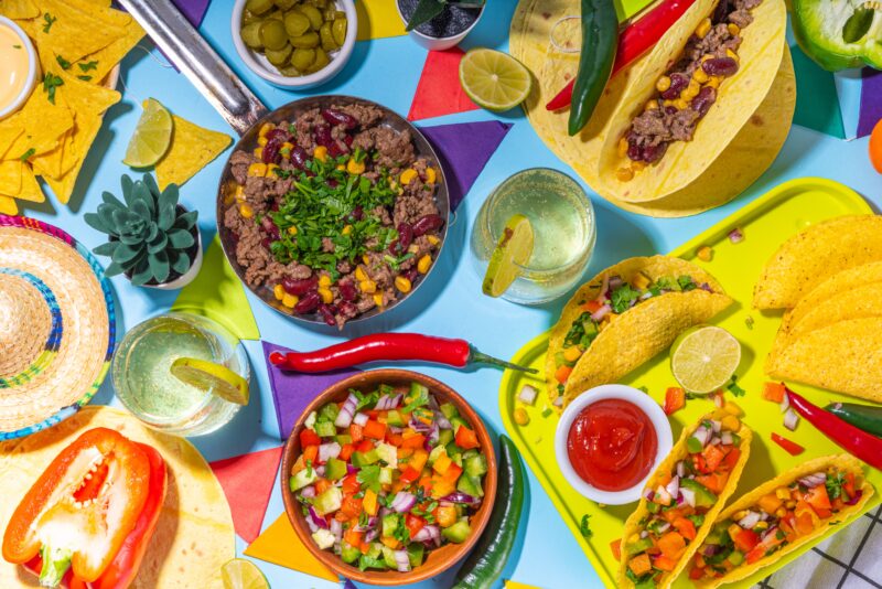 Alt text: Colorful top-down view of Mexican-style food including tacos, tortilla chips, ground beef with beans and corn, fresh salsa, lime wedges, and chili peppers, arranged on a vibrant table with drinks and a small cactus.