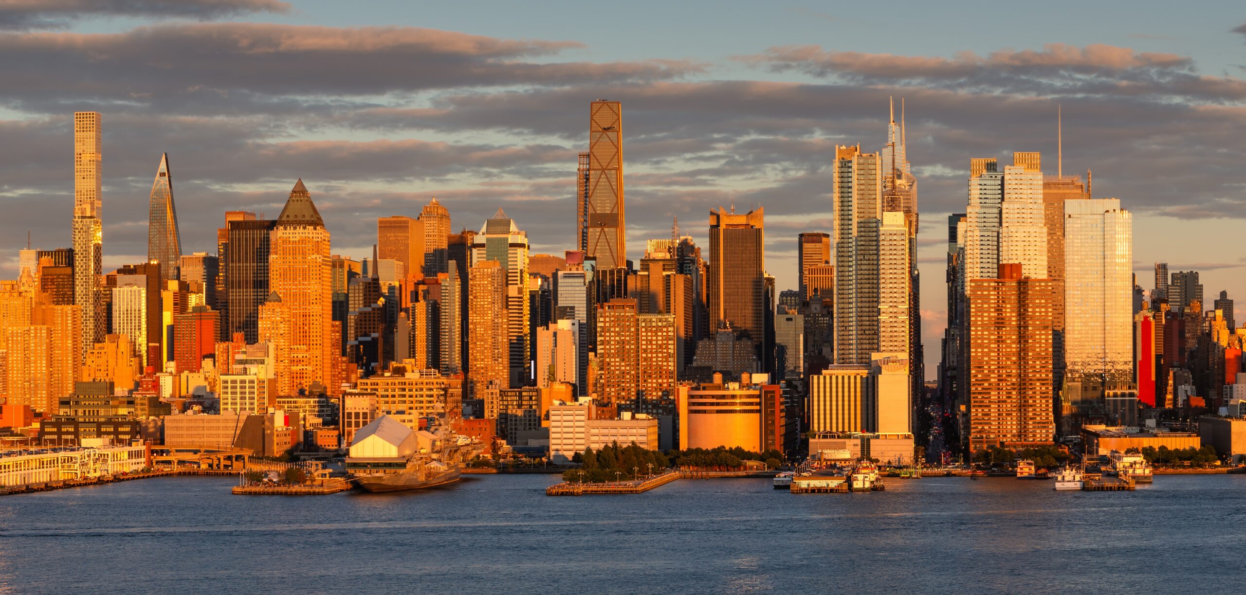 Golden hour view of the Manhattan skyline from the waterfront, with Midtown skyscrapers glowing in warm sunset light above the Hudson River.