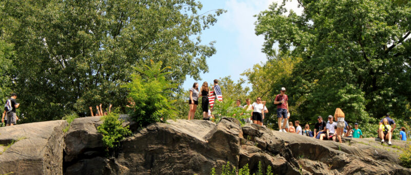 Visitors gather on large rock formations in Central Park, enjoying scenic views and sunny weather while surrounded by lush trees and greenery.