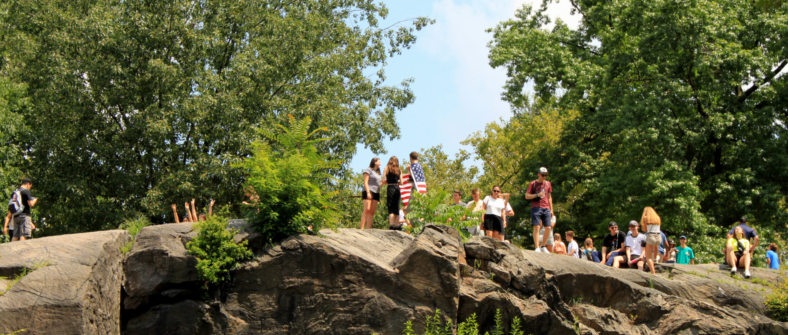 Visitors gather on large rock formations in Central Park, enjoying scenic views and sunny weather while surrounded by lush trees and greenery.