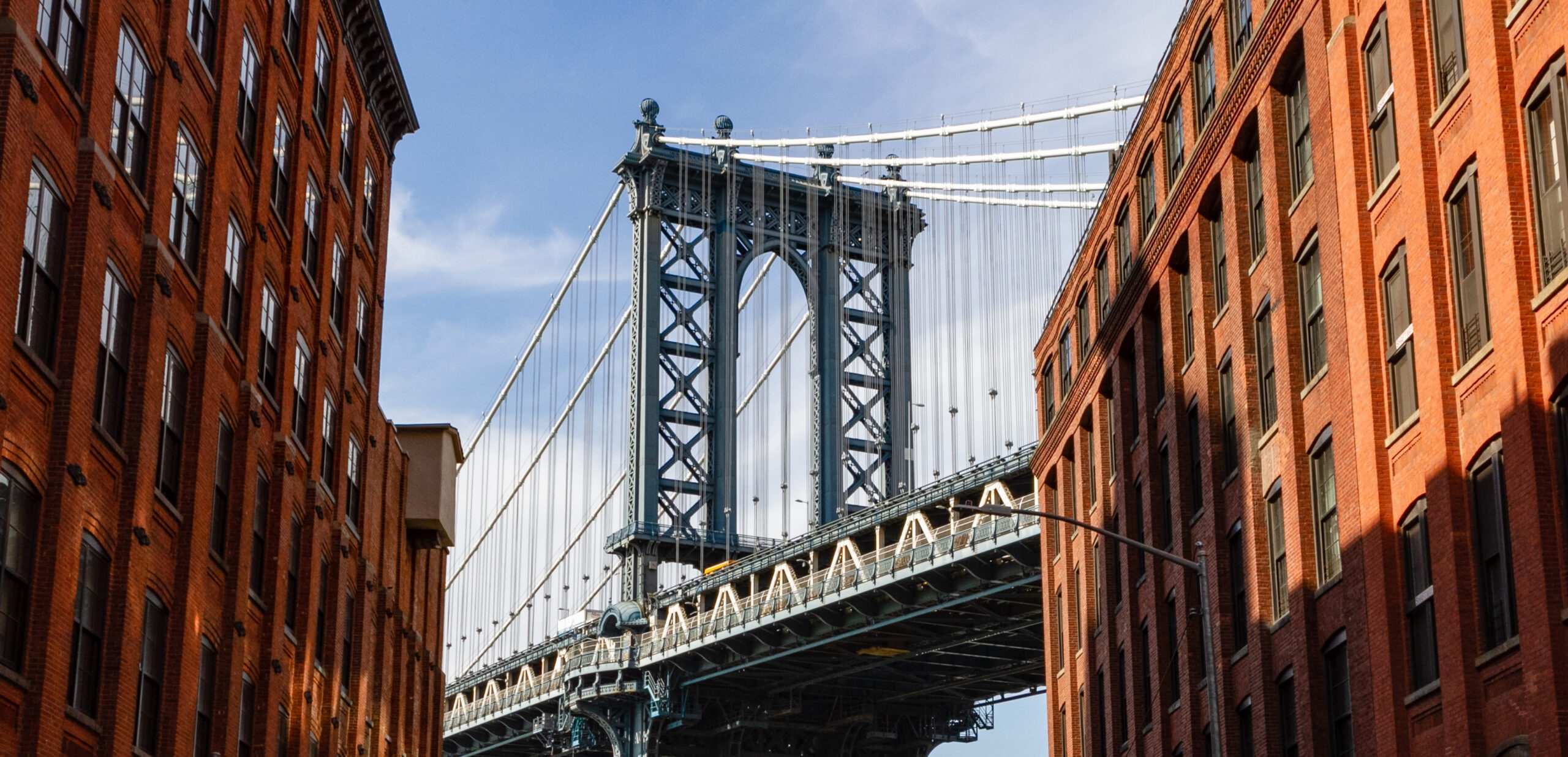 View of the Manhattan Bridge from Washington Street in DUMBO, Brooklyn, with historic warehouse buildings and the Empire State Building visible in the distance.