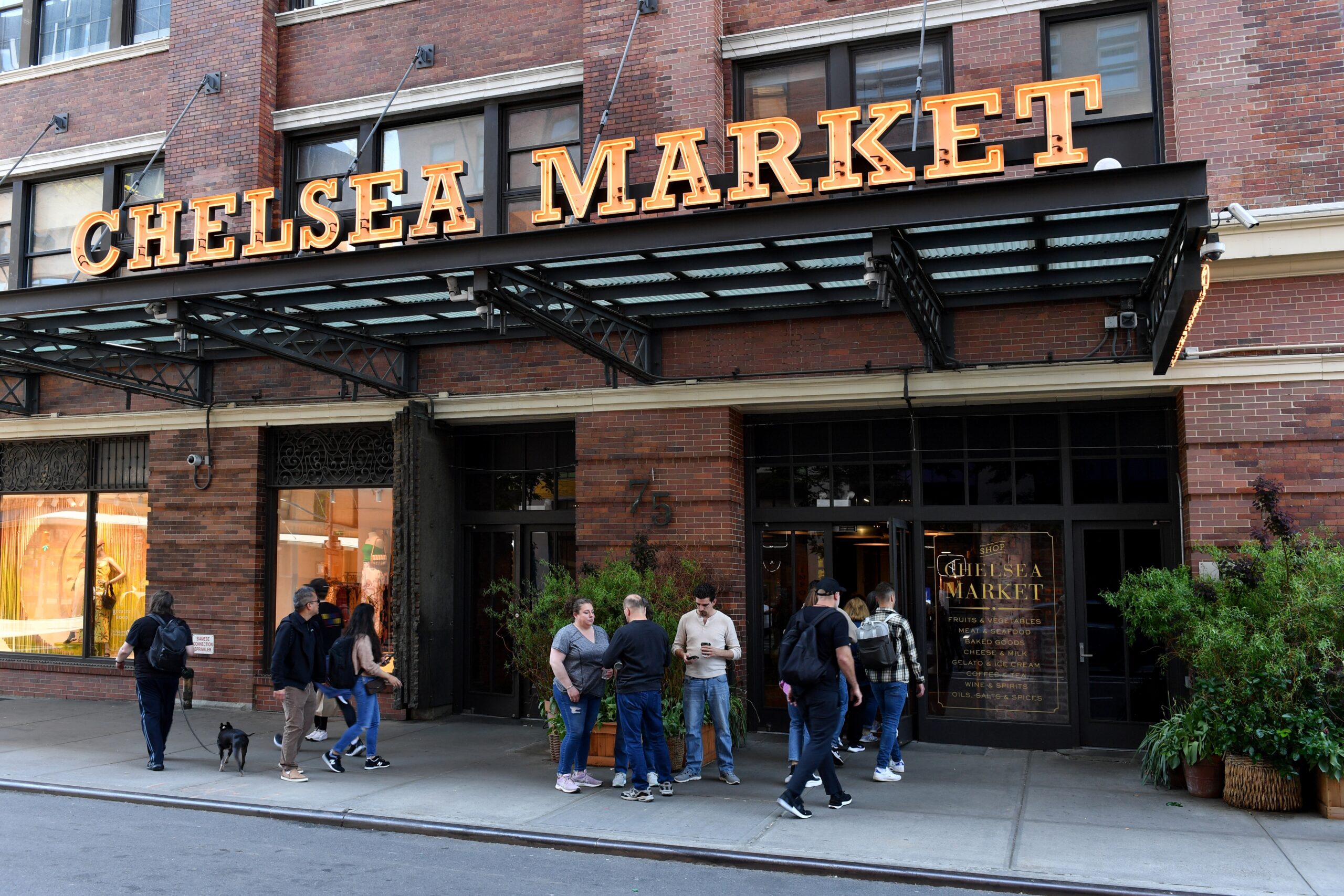 Chelsea Market exterior entrance on 9th Avenue, with its illuminated sign above the brick façade and visitors gathered outside.