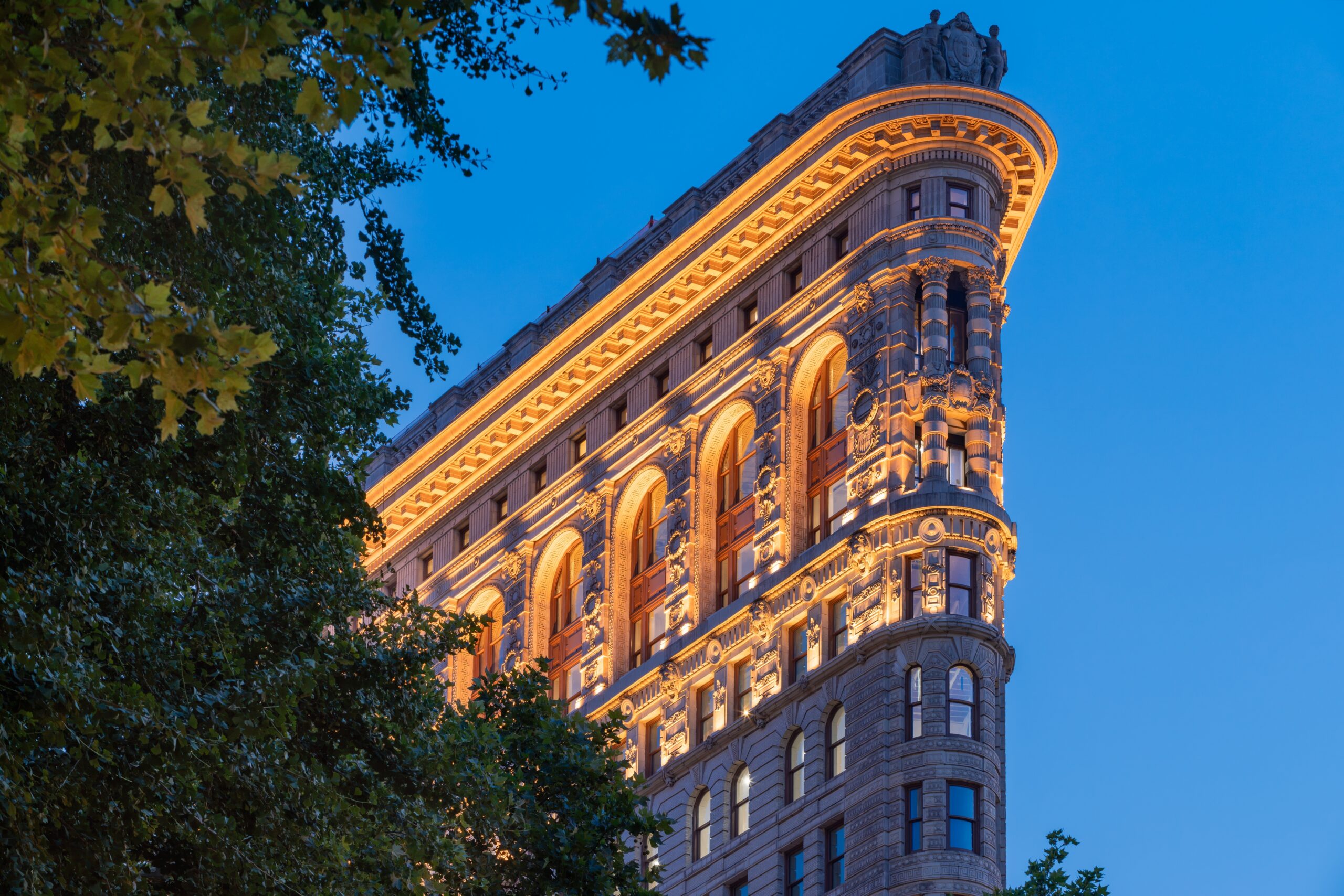 The Flatiron Building is illuminated at dusk, viewed from Madison Square Park with trees framing its ornate triangular facade against a blue evening sky.