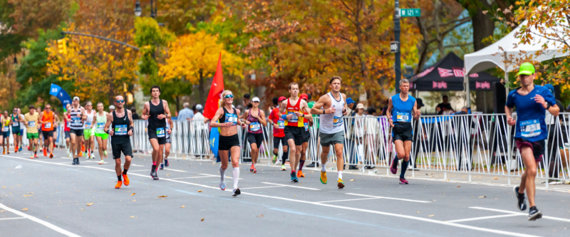 Runners compete in the New York City Marathon on a tree-lined Manhattan street, surrounded by colorful autumn foliage and race barriers.