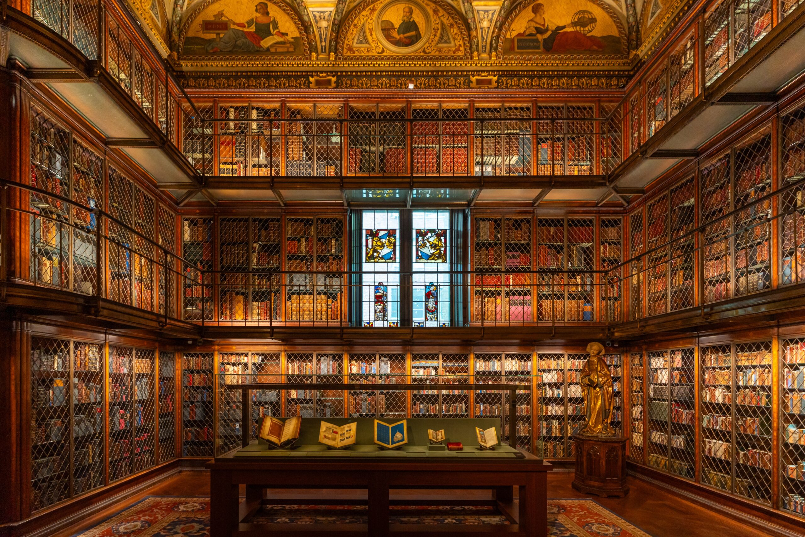 The Morgan Library & Museum’s historic library interior, featuring ornate wood-paneled bookshelves, intricate metal grilles, stained-glass windows, gilded ceiling decorations, and rare books displayed in the center.