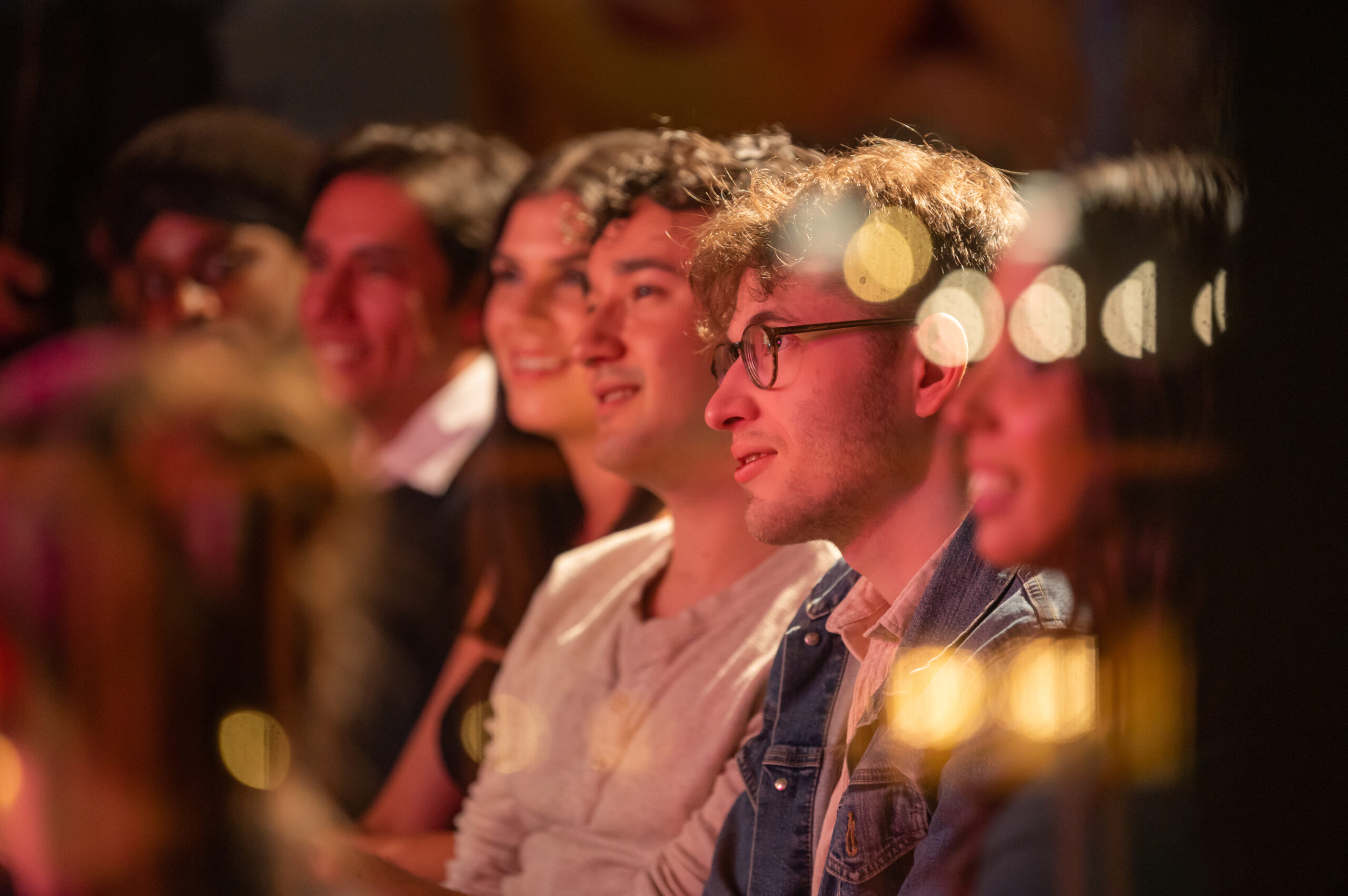 A group of people, mostly young adults, are sitting in a dimly lit venue with bokeh-style lights adding warmth to the scene, focused on something off-camera.