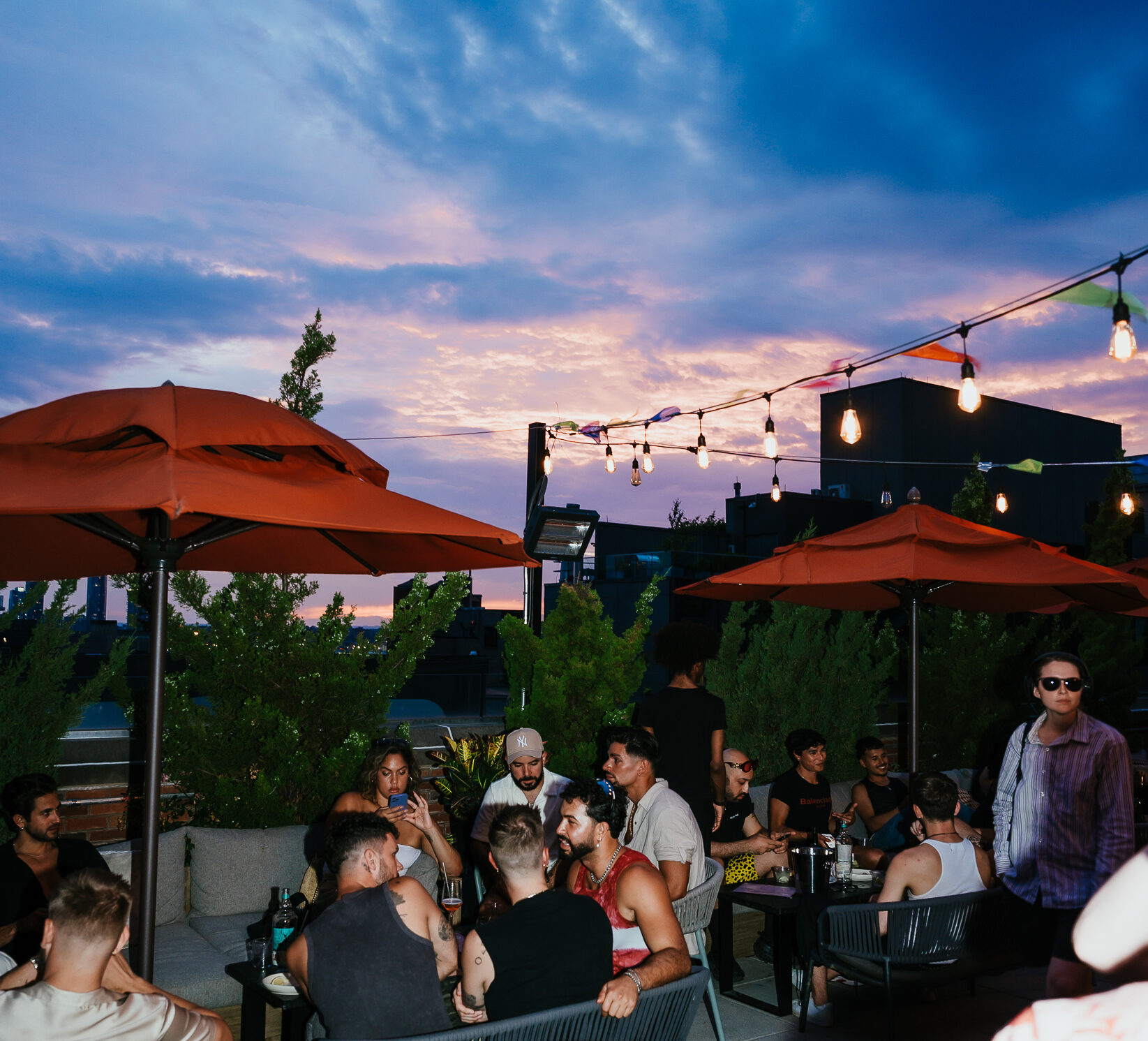 A group of people enjoy an evening at Arlo SoHogathering on a rooftop patio with string lights and orange umbrellas, against a backdrop of a colorful sunset sky.
