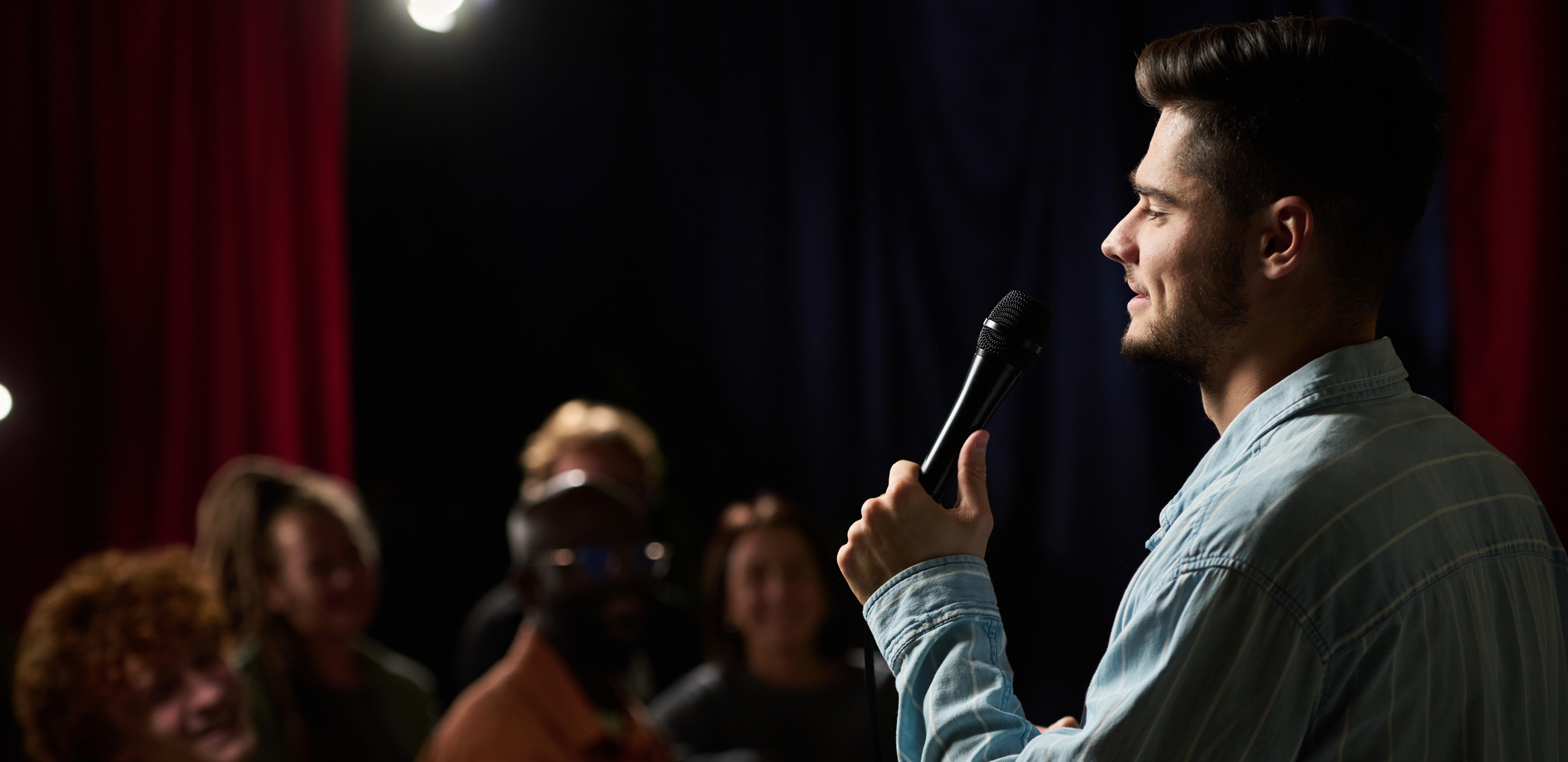 A comedian smiles while performing a stand-up set into a microphone for a laughing audience in a dimly lit club.