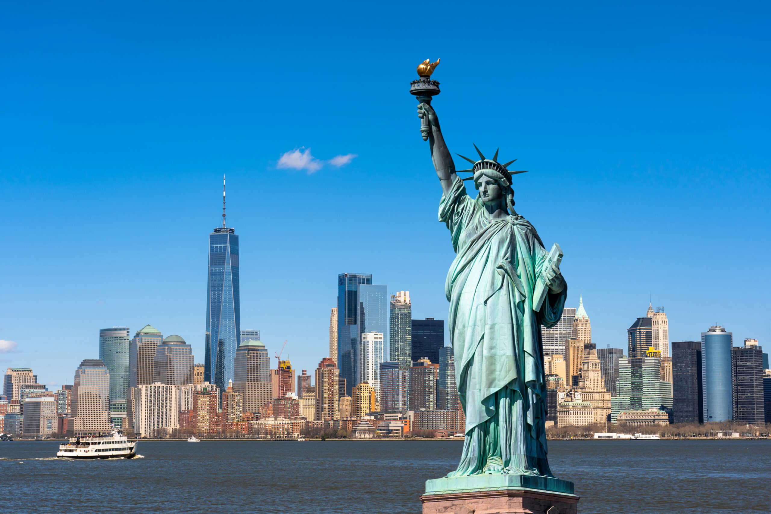 The Statue of Liberty standing prominently in New York Harbor with a Liberty Cruise ferry and the Manhattan skyline in the background.