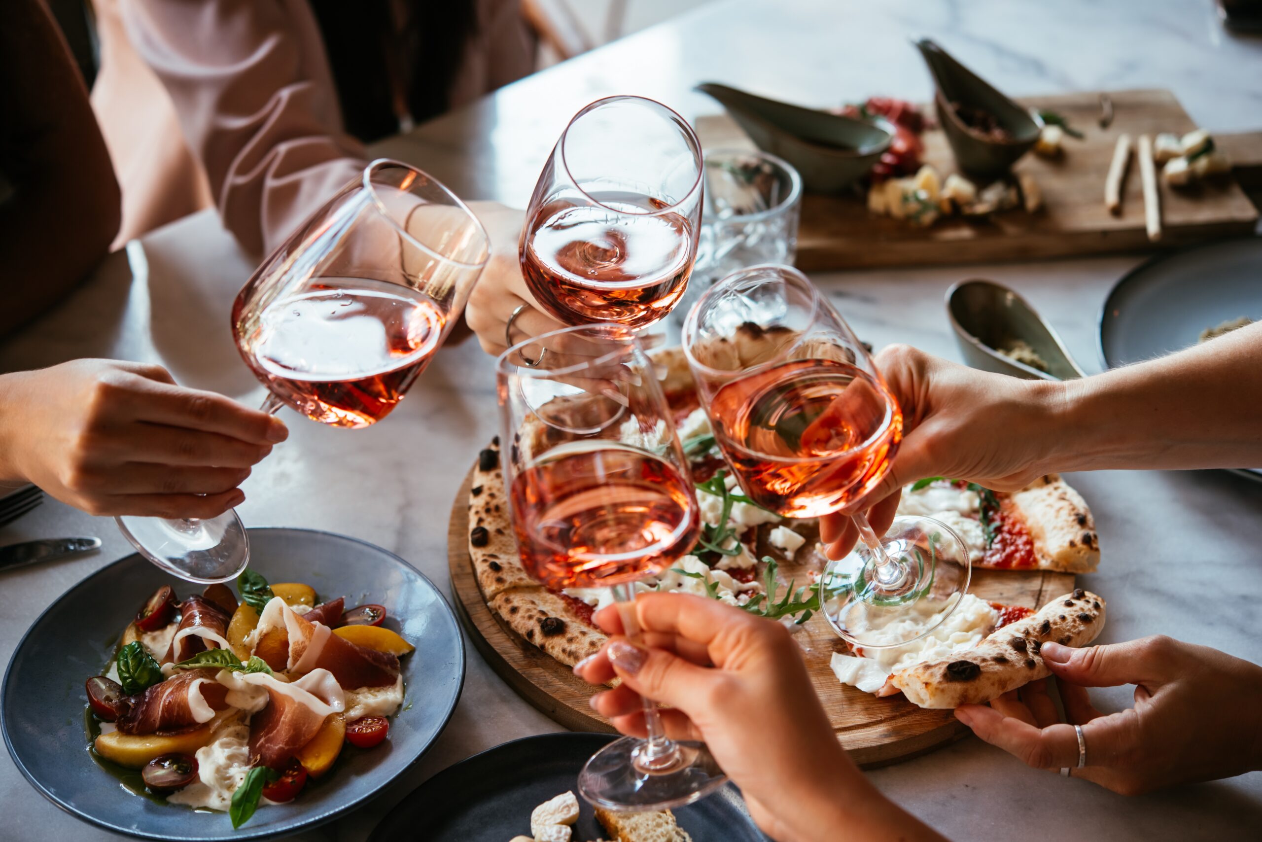Friends toast with glasses of rosé wine over a table spread with Neapolitan pizza and an antipasto salad in a trending restaurant