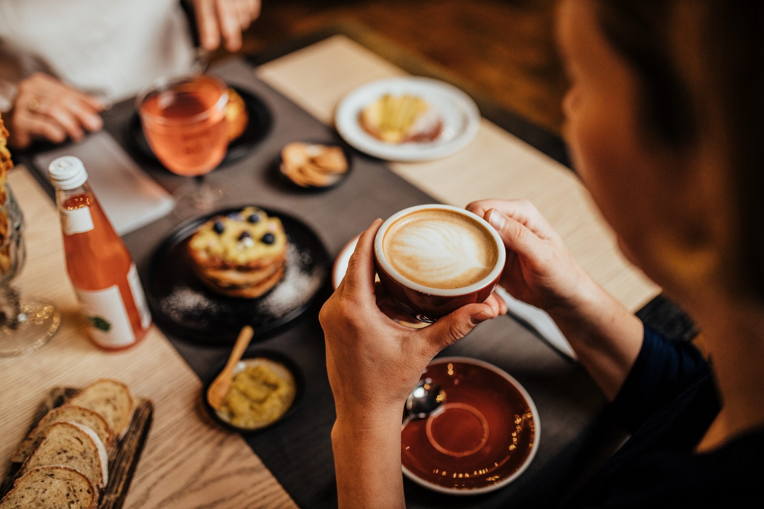 A close-up, first-person view of a person holding a warm latte with heart-shaped latte art in a red ceramic cup in one of the SoHo’s Cafés