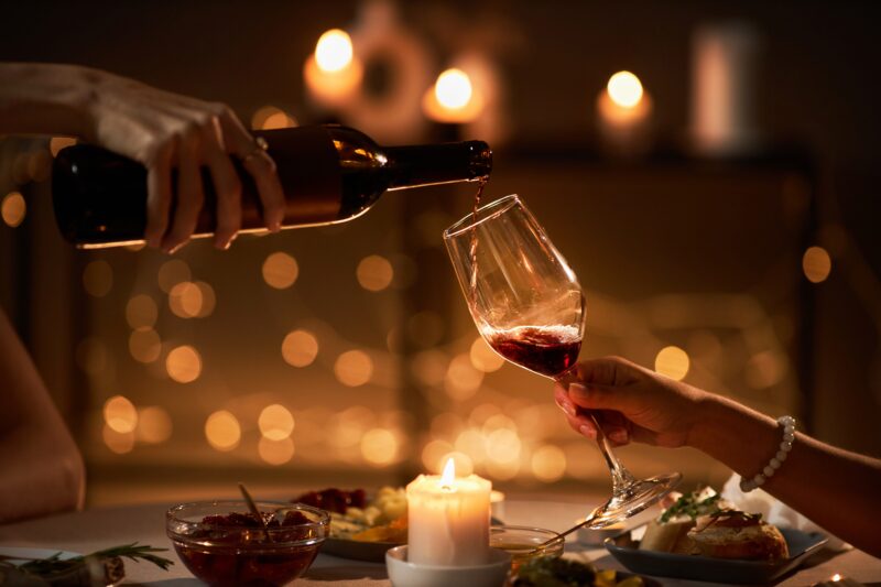 Gemini said A close-up, atmospheric shot of a romantic candlelit dinner. One person pours red wine from a dark bottle into a glass held by another person.