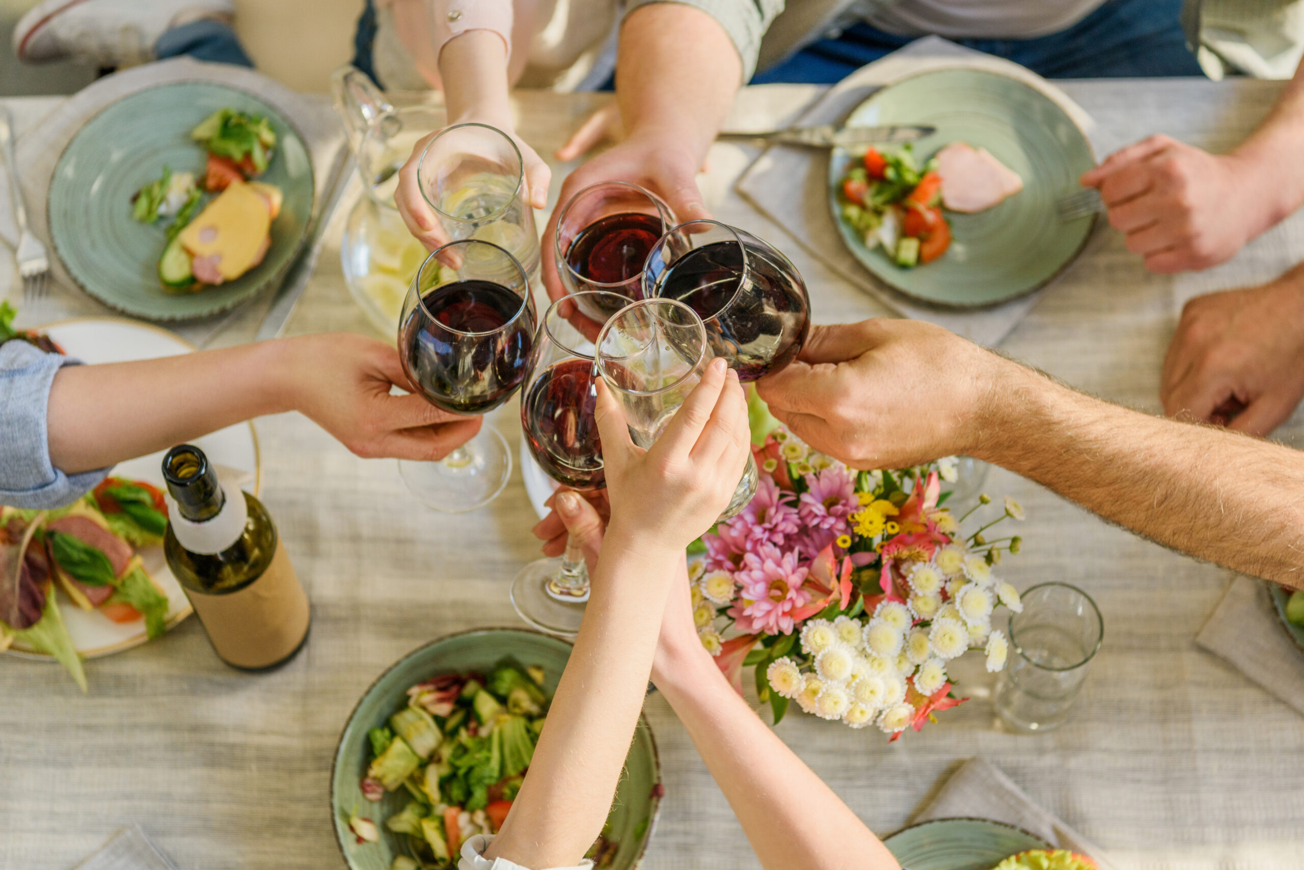 An overhead view of several people raising their wine and water glasses in a toast over a rustic dinner table set with fresh salads and a vibrant bouquet of pink and white flowers at Antique Garage.