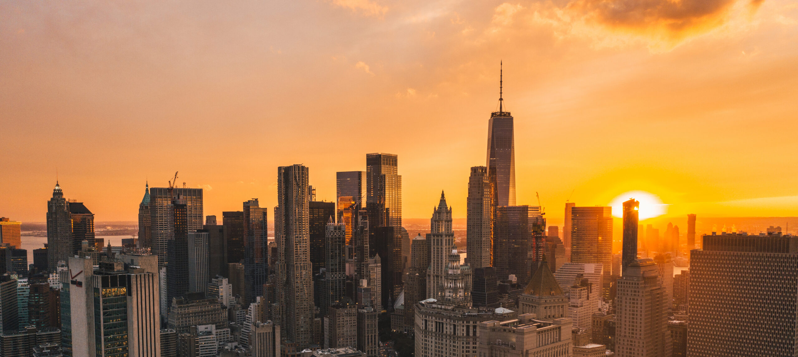 An aerial, wide-angle view of the Lower Manhattan skyline during Manhattanhenge, featuring One World Trade Center prominently in the center. The sun is perfectly aligned with the city's east-west grid, positioned low on the horizon to the right and glowing intensely. This alignment creates a dramatic golden-orange light that floods the canyons between skyscrapers and illuminates the hazy sky.