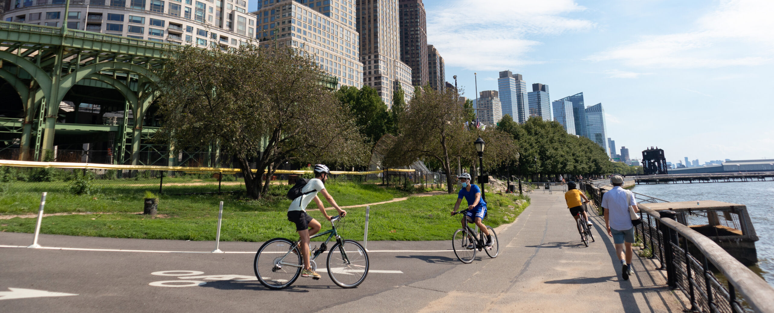 Cyclists ride along the Hudson River Greenway near Battery Park, with waterfront views, green park space, and Lower Manhattan skyline buildings in the background.