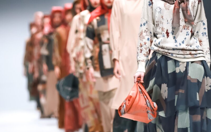 Models walk down the runway during New York Fashion Week wearing modest fashion looks with layered garments, floral prints, and colorful headscarves, with the front model carrying a coral handbag while others appear softly blurred in the background.