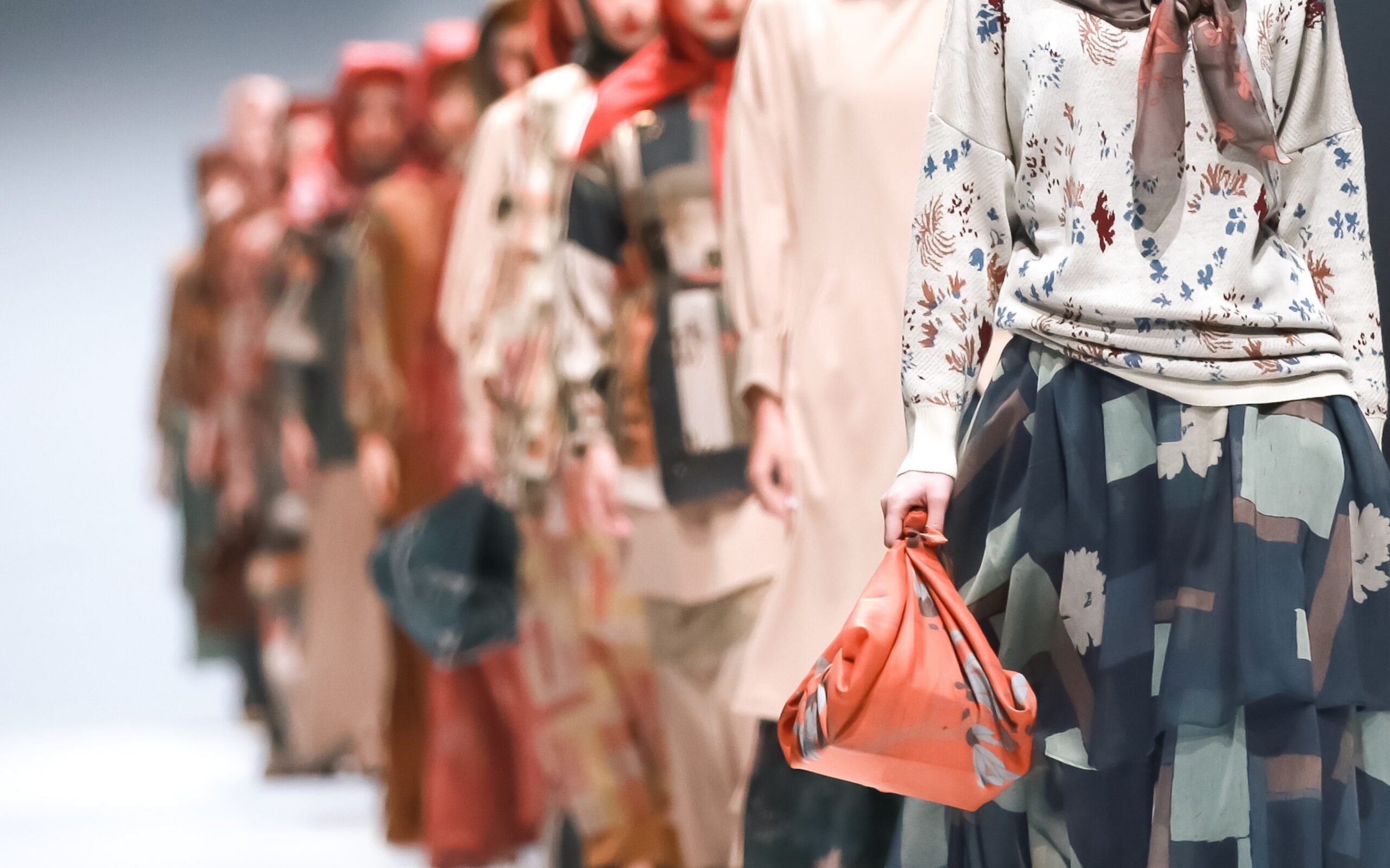 Models walk down the runway during New York Fashion Week wearing modest fashion looks with layered garments, floral prints, and colorful headscarves, with the front model carrying a coral handbag while others appear softly blurred in the background.