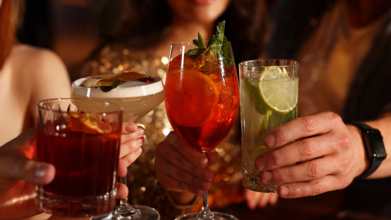 Four people raise colorful cocktails for a toast during a night out, with drinks garnished with citrus and fresh mint.