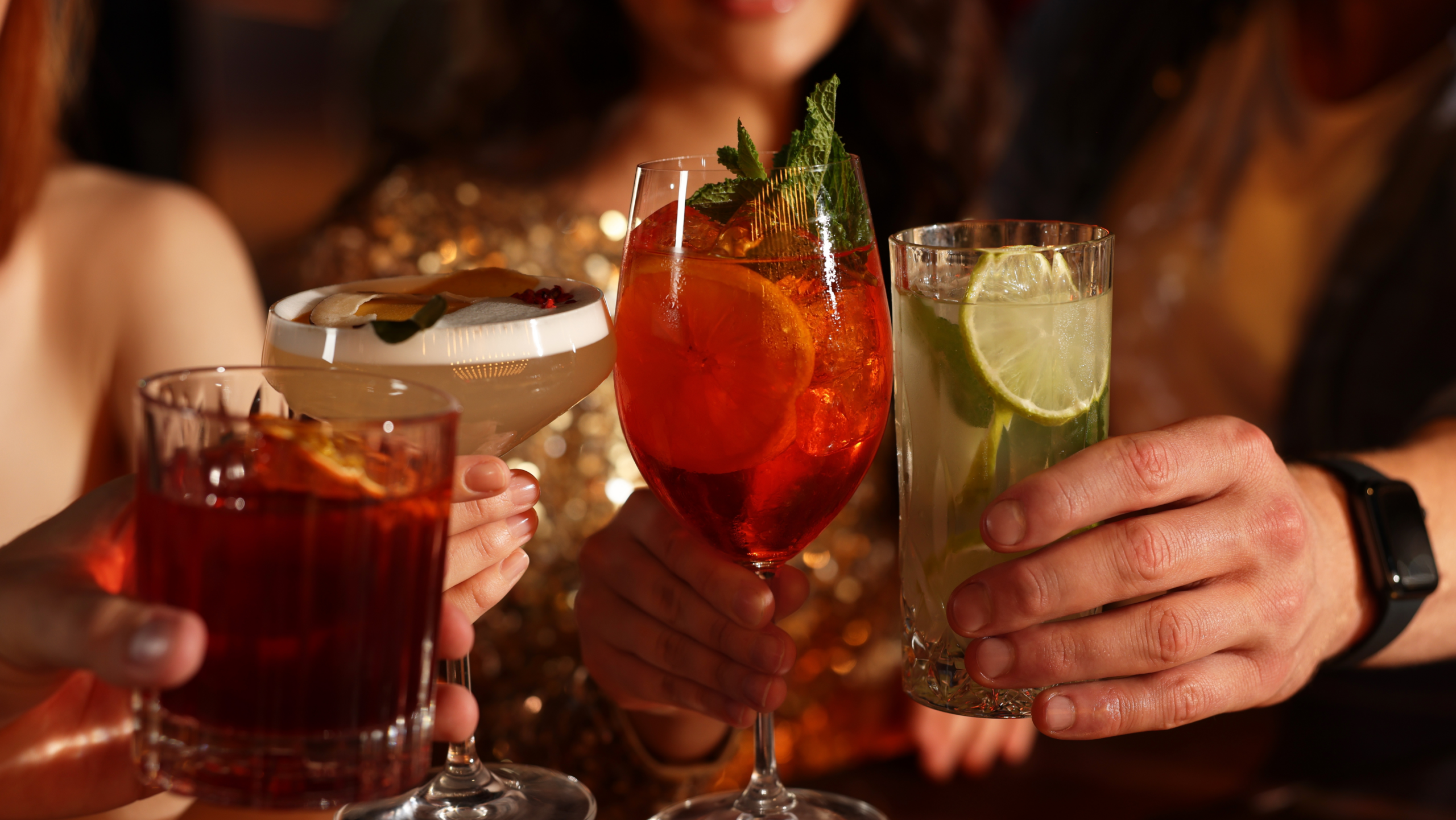 Four people raise colorful cocktails for a toast during a night out, with drinks garnished with citrus and fresh mint.